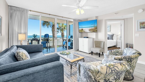A modern living room with a sofa, armchair, and a coffee table. There is a large window with a view of palm trees and the ocean. - 🌴Island Princess 203 | Gulf Views | Pool + Hot Tub🌴 (Fort Walton Beach)