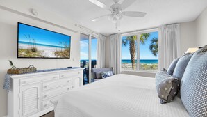 A bedroom with a bed, a TV, a ceiling fan, a dresser, and a large window with a view of the beach. - 🌴Island Princess 203 | Gulf Views | Pool + Hot Tub🌴 (Fort Walton Beach)