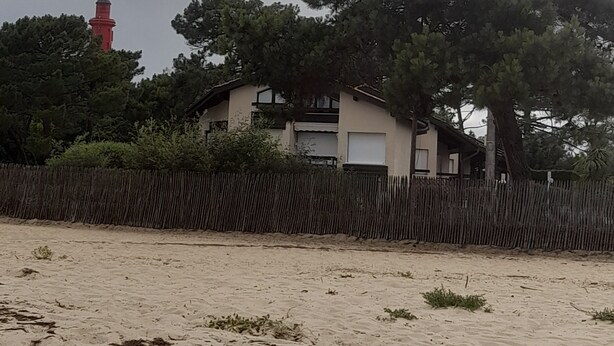 A coastal house with a sandy foreground, a wooden fence, and a red lighthouse in the background. - Apartment in Cap Ferret, across from Phare Beach (Lège-Cap-Ferret)