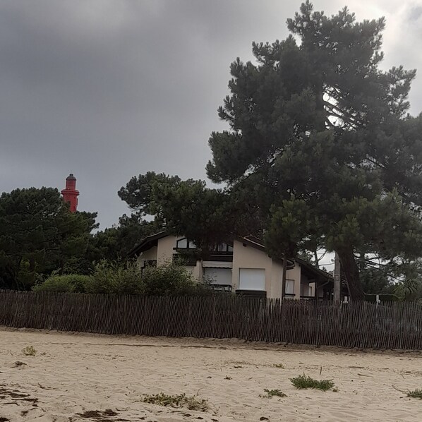 A coastal house with a sandy foreground, a wooden fence, and a red lighthouse in the background. - Apartment in Cap Ferret, across from Phare Beach (Lège-Cap-Ferret)