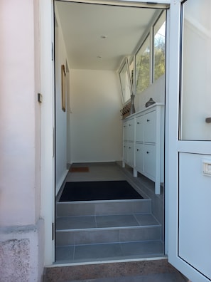 A narrow hallway with a set of stairs leading to a room. There is a white door on the right, a white cabinet with multiple drawers, and a black mat at the bottom of the stairs.