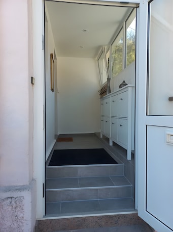 A narrow hallway with a set of stairs leading to a room. There is a white door on the right, a white cabinet with multiple drawers, and a black mat at the bottom of the stairs.. Maison La Mirabelle – Peaceful Retreat at the Edge of the Forest