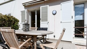 Een terras buiten met een ronde tafel en houten stoelen, een barbecue, en een wit gebouw met luiken.
