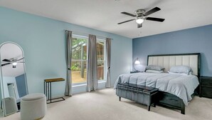 Une chambre à coucher comprenant un lit, un ventilateur de plafond, une table de chevet, une commode, un miroir et une fenêtre avec des rideaux.