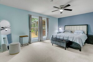 Un dormitorio con cama, ventilador de techo, mesita de noche, espejo y ventana con cortinas.