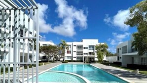 A swimming pool with a clear blue sky, modern buildings, and a gated area. - Apartment with private access to the beach (Punta Cana)