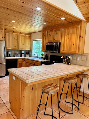 A kitchen with wooden cabinets, a stainless steel refrigerator, a microwave, and a large island with stools. - A Cozy Mountain Getaway in Arnold! (Arnold)