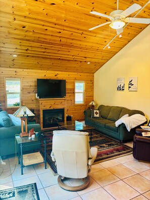 A cozy living room with a fireplace, a television, a green sofa, a leather armchair, and a ceiling fan. - A Cozy Mountain Getaway in Arnold! (Arnold)