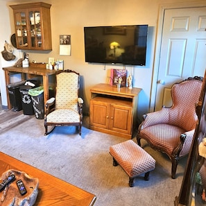 A living room with a television, wooden cabinet, patterned armchair, ottoman, and a small table with remotes.