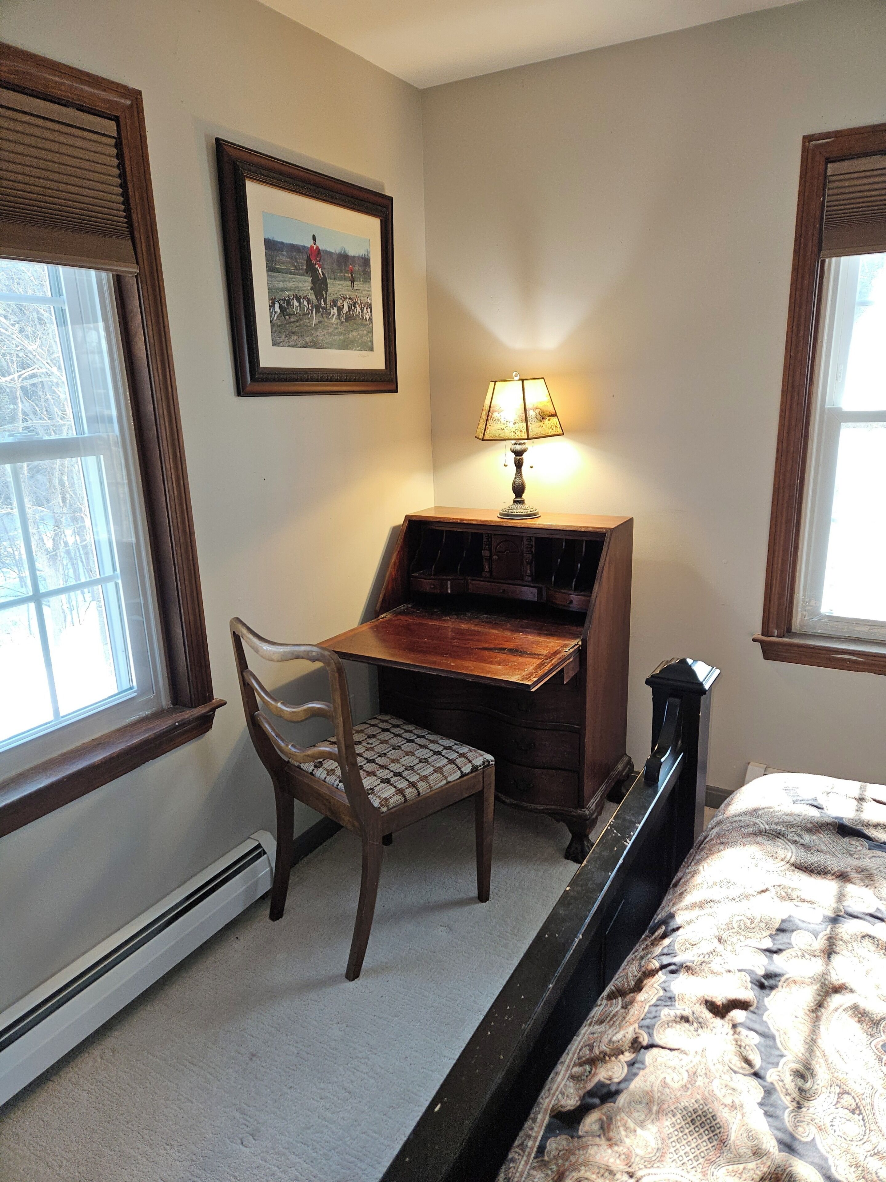 A bedroom with a bed, a desk with a chair, a lamp, a framed picture on the wall, and a window.