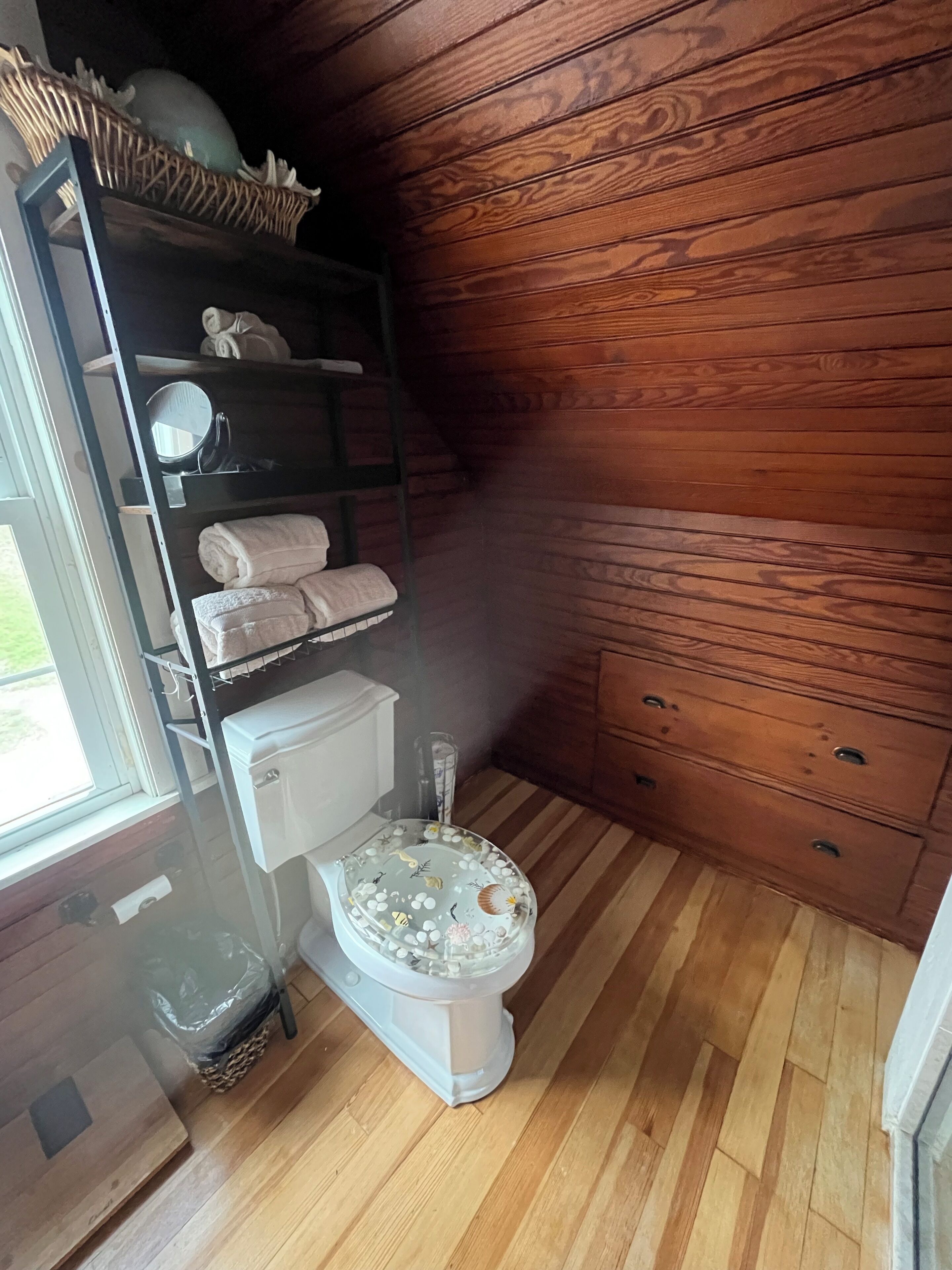 A bathroom with a wooden floor, a white toilet, a shelf with towels, and a window.