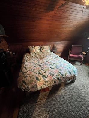 A bedroom with a wooden ceiling, a floral bedspread, a chair, and a nightstand with a lamp.