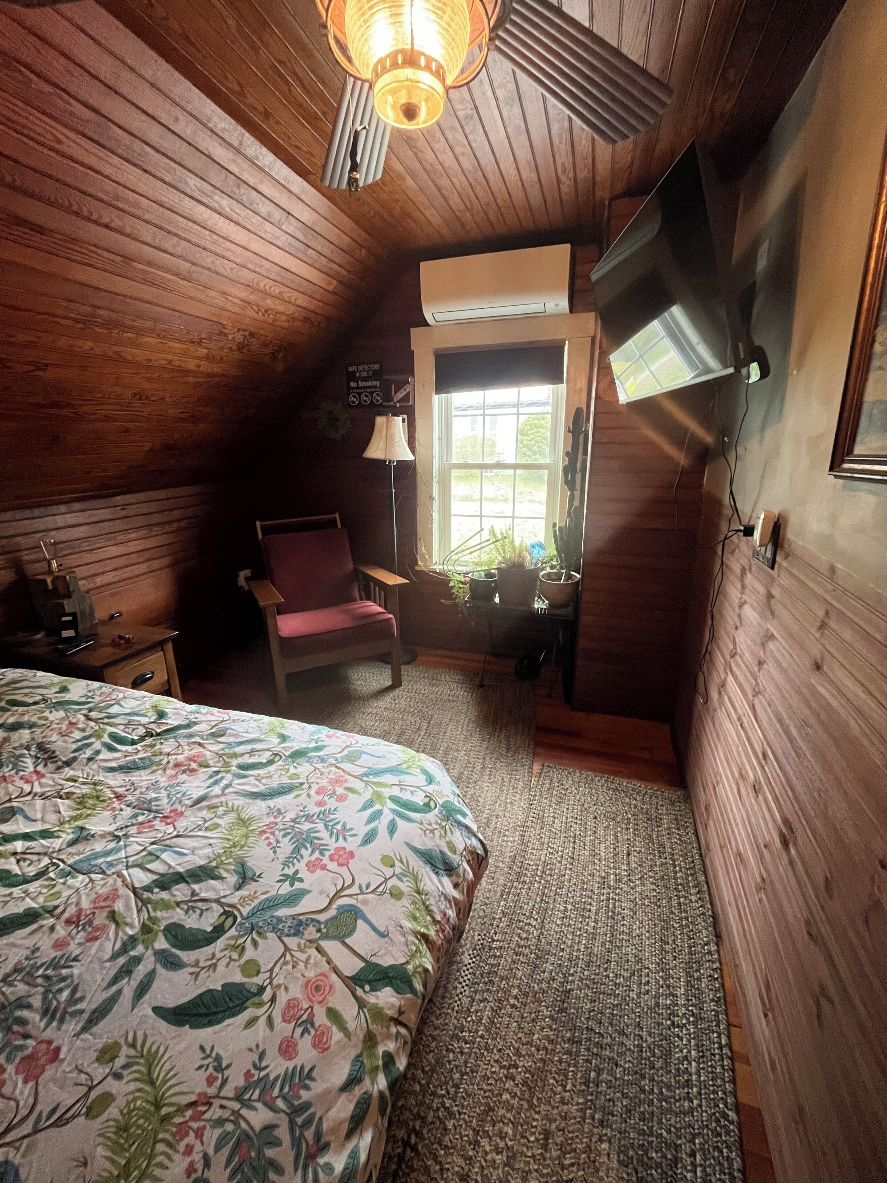 A room with wooden walls, a bed with a floral bedspread, a chair, a lamp, and a window with plants.