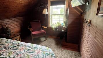 A room with wooden walls, a bed with a floral bedspread, a chair, a lamp, and a window with plants.
