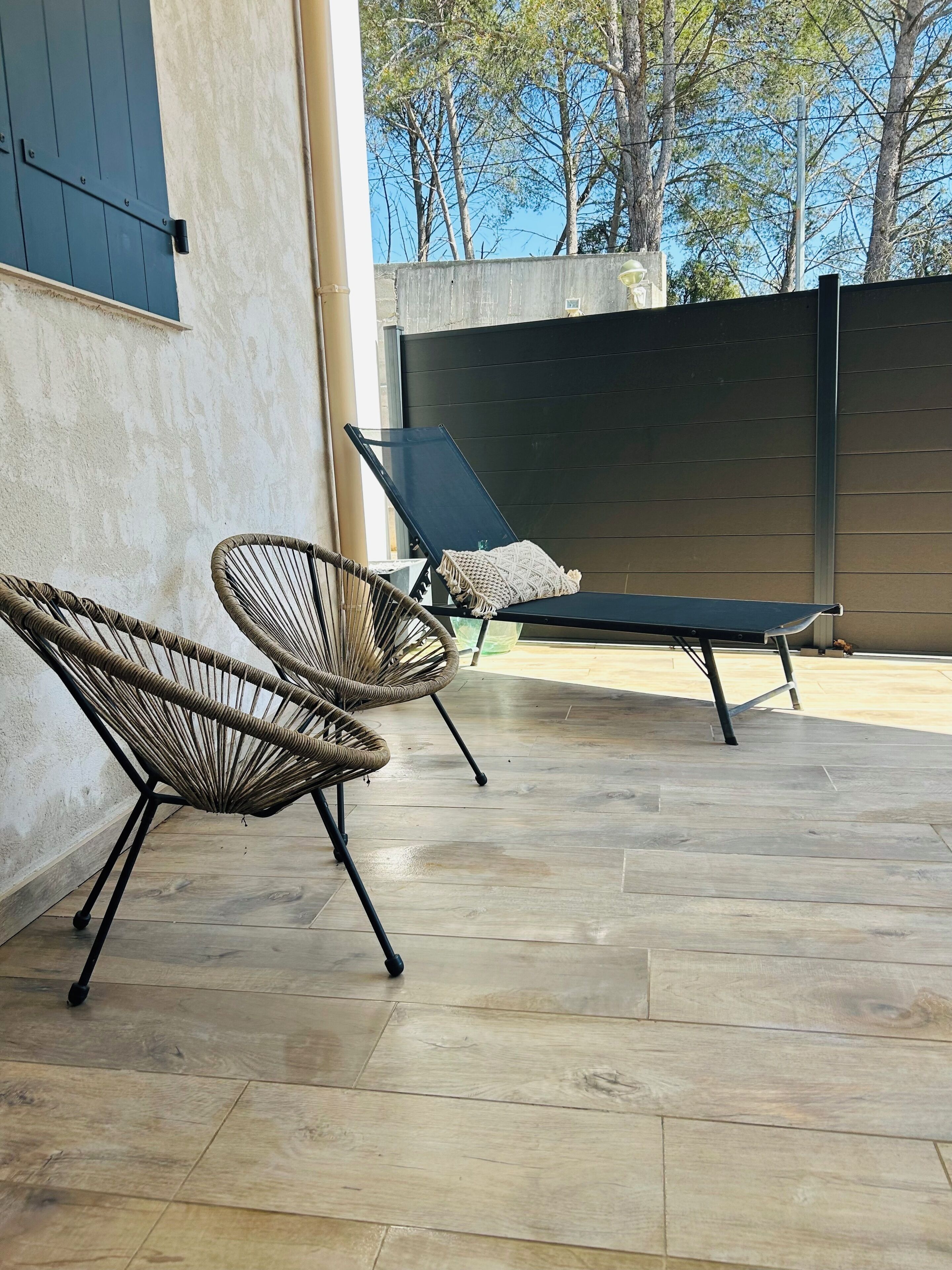A patio with two wicker chairs and a lounge chair, a tiled floor, and a view of trees.