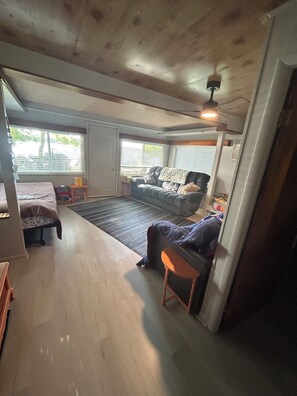Ein geräumiges Zimmer mit großem Fenster, einem Sofa, einem Sessel und einem Deckenventilator.