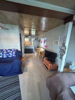 Ein kleines Zimmer mit einem Bett, einem Fernseher, einem kleinen Esstisch und einer Küchenzeile im Hintergrund.