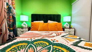 Un dormitorio con una pared verde, una cama con colcha estampada, dos lámparas de noche y una mesita de noche con un objeto decorativo.