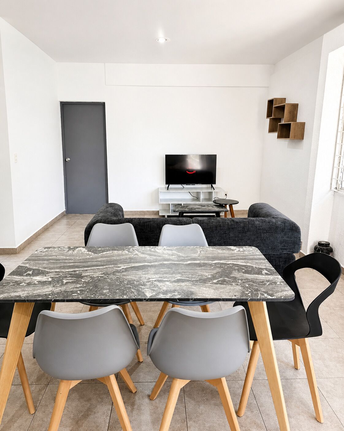 Een moderne woonkamer met een marmeren eettafel, grijze stoelen, een bank, een televisie en een deur.