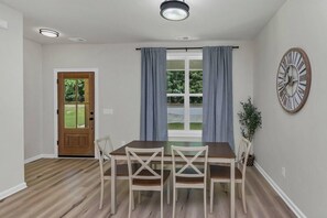 Un coin repas avec une table et des chaises en bois, une fenêtre avec des rideaux, une horloge au mur et une porte donnant sur l’extérieur.