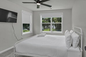 Une chambre à coucher avec un lit, des oreillers blancs, un ventilateur de plafond et deux fenêtres donnant sur un paysage verdoyant.