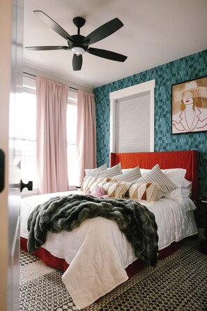 Une chambre à coucher avec un lit, un ventilateur de plafond, une fenêtre avec des rideaux et un mur orné d’un motif géométrique.