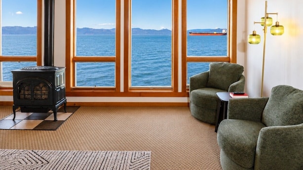 A cozy room with a large window, a wood-burning stove, and a green armchair. - Pier 39 Over the water full River View + Fireplace (Astoria)