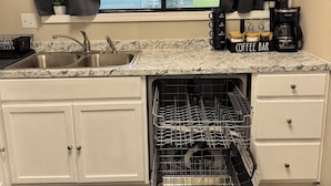 Una cocina con lavavajillas, cafetera y ventana con cortinas.