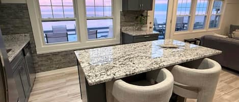 A kitchen with a granite countertop, two barstools, a stone backsplash, and large windows overlooking a body of water.