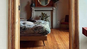 Un dormitorio con piso de madera, una cama con ropa de cama estampada, una lámpara colgante, un espejo sobre la chimenea y un marco de puerta.