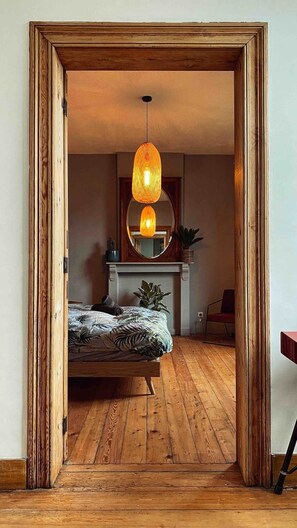 Ein Schlafzimmer mit Holzboden, einem Bett mit gemusterten Textilien, einer Hängeleuchte, einem Spiegel über einem Kamin und einem Türrahmen.