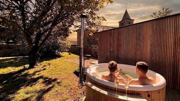 Una bañera de madera con dos personas, un árbol y un muro de piedra al fondo.