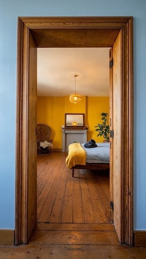 Une pièce avec un plancher en bois, un lit recouvert d’une couverture jaune, un luminaire suspendu et une plante.