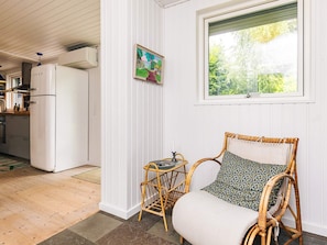 Una habitación con una silla de mimbre, una mesita, una ventana con vistas a zonas verdes, una zona de cocina con refrigerador e imagen colgada en la pared.
