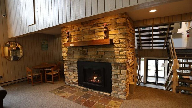 Una chimenea de piedra con repisa de madera, un comedor con sillas y una escalera que sube a un nivel superior.