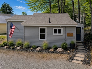 A small cabin with a flag, a welcome sign, and a gravel path leading to the door.