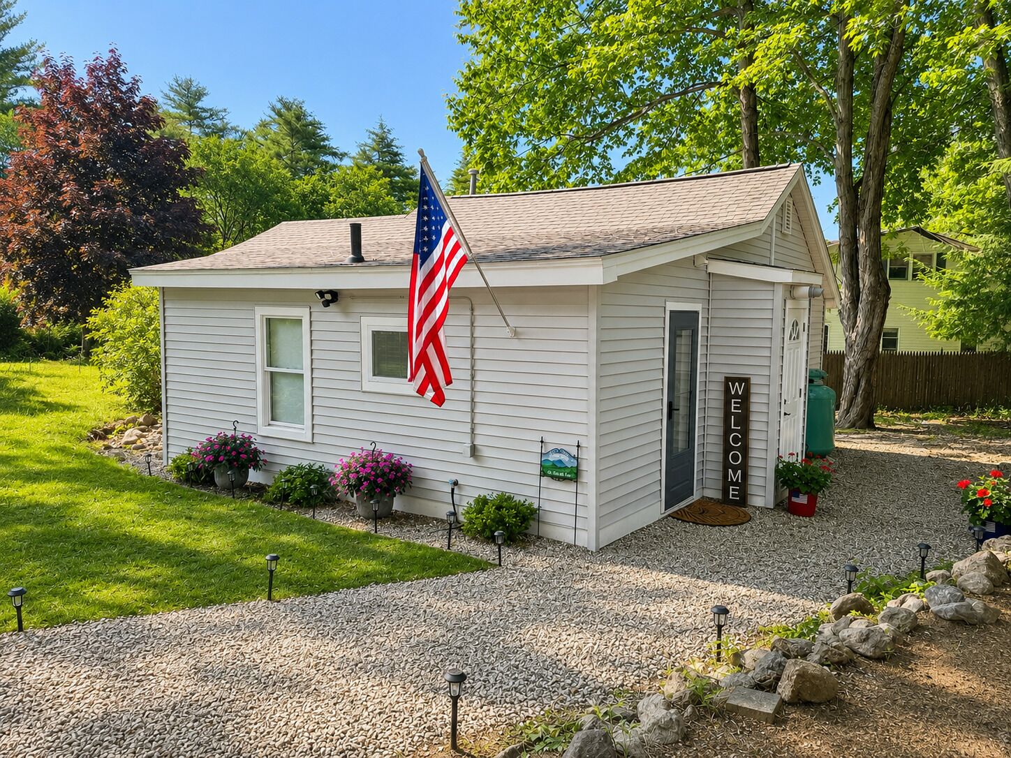 A small white house with a flag, a welcome sign, and potted plants.