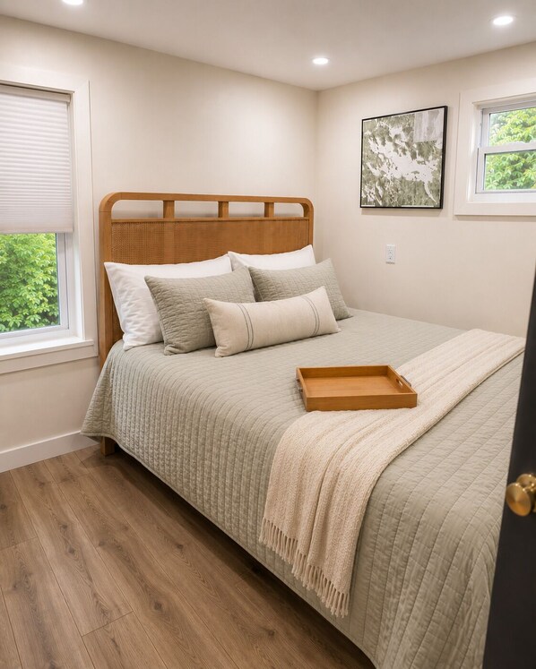 A bedroom with a wooden headboard, a bed with a beige quilt, pillows, a bedside table with a tray, and a window with blinds. - Charming 3BR Home on Paugus Bay with Shared Beach (Laconia)