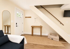 Una sala de estar con un sofá, una alfombra, una cama para perro, una mesa de madera, un perchero, una puerta y una escalera.