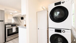A laundry room with stacked washers and dryers, a white door, and a kitchen area with a stove and oven. - Lake Erie Retreat (Dunkirk)