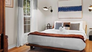 Ein Schlafzimmer mit Bett, einem Fenster mit Vorhängen, einer Wandlampe und einem Gemälde.