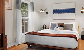 Ein Schlafzimmer mit Bett, einem Fenster mit Vorhängen, einer Wandlampe und einem Gemälde.