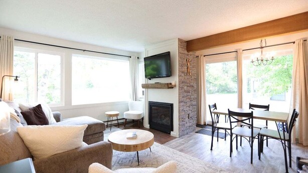 A living room with a fireplace, a television, a dining area, and large windows. - Perfect Location at this Waterville Condo! (Waterville Valley)