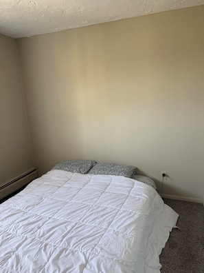 Une chambre à coucher avec un couvre-lit blanc, des coussins à motifs, une prise électrique visible et un sol recouvert de moquette.