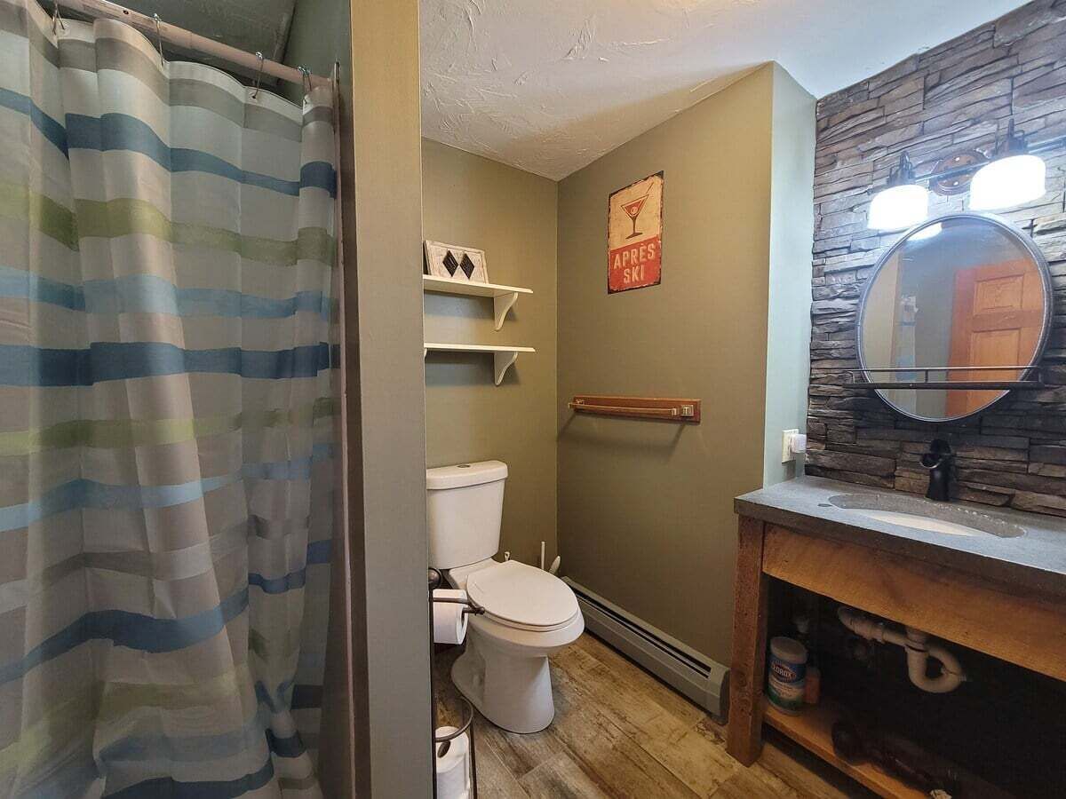 A bathroom with a striped shower curtain, a toilet, a vanity with a mirror, and a wall-mounted shelf.