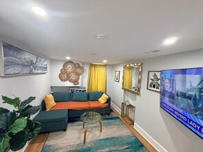 A living room with a sectional sofa, a glass coffee table, a flat-screen TV, and a large plant. - Charming 3-bedroom house in Washington with cool AC breeze (Washington)