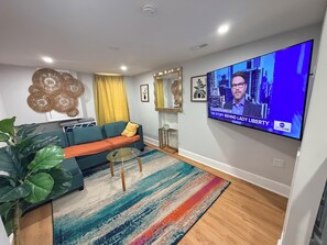Ein modernes Wohnzimmer mit Sofa, Couchtisch, Fernseher, der eine Nachrichtensendung zeigt, und einer großen Pflanze.