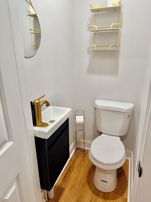 Une petite salle de bain avec des toilettes blanches, un lavabo avec un robinet doré, un meuble noir, un miroir et une étagère blanche agrémentée de détails dorés.