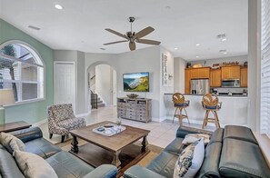 A living room with a sofa, armchairs, a coffee table, a TV, and a kitchen area with wooden cabinets and a refrigerator.