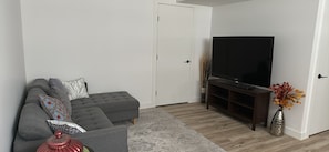 A living room with a grey sofa, a flat-screen TV, a wooden entertainment unit, and a vase with flowers. - Cozy basement suit in Rosewood near Costco, Saskatoon (Saskatoon, SK)